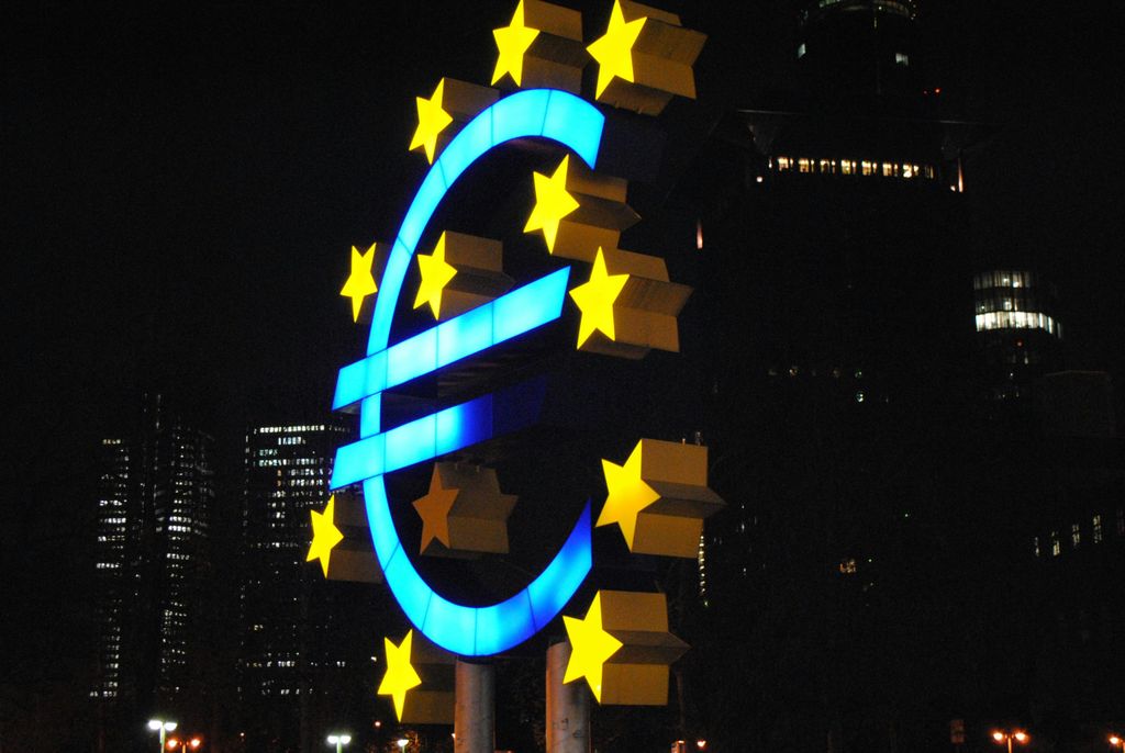 A neon blue and yellow euro sign with white stars at night amidst dark bulidings in Frankfurt, Germany.