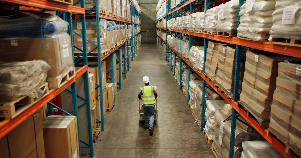 A man walks down a warehouse corridor in a hard hat and high-vis jacket.