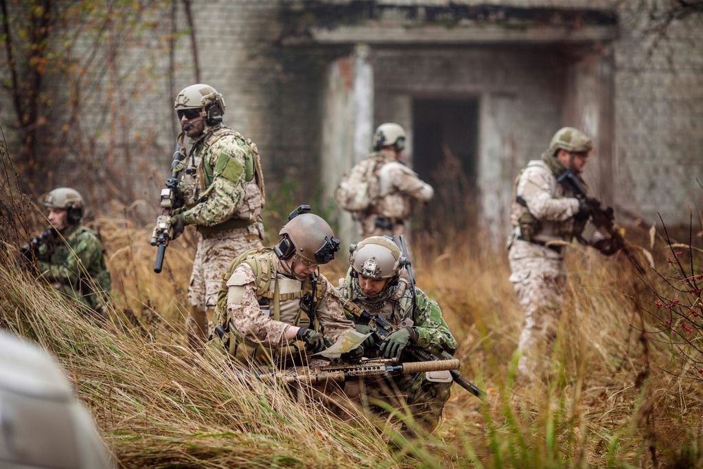 Military personnel in a simulation. Four armed soldiers in fatigues stand watch over two soldiers examining a map in a wooded area.